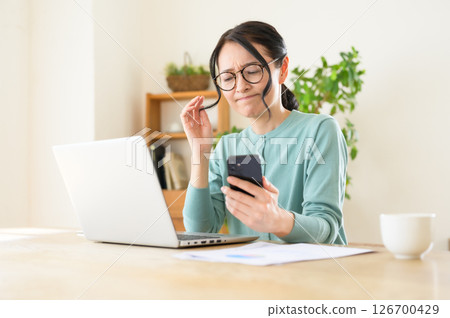 A middle-aged woman is worried while looking at a computer with a smartphone in one hand A middle-aged woman is worried while looking at a computer with a smartphone in one hand 126700429