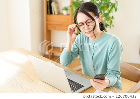 A middle-aged woman is worried while looking at a computer with a smartphone in one hand A middle-aged woman is worried while looking at a computer with a smartphone in one hand 126700432
