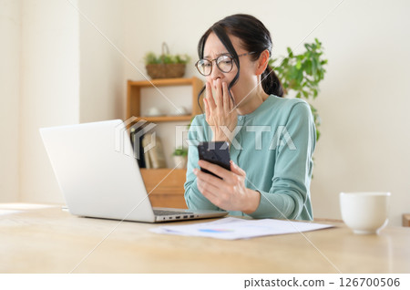 一位中年婦女一手拿著智慧型手機，一邊看著電腦，一邊感到驚訝 126700506