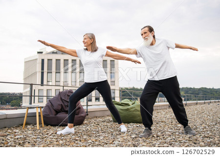 Elderly man and woman performing yoga together on rooftop in warrior pose, promoting well-being and fitness Elderly man and woman performing yoga together on rooftop in warrior pose, promoting well-being and fitness 126702529
