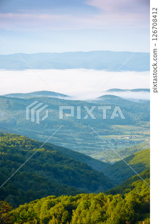 mountainous countryside landscape with fog in the distant valley. mist rolling among hills in morning light. stunning view of carpathians in summer. clouds on the blue sky. amazing dale 126708412