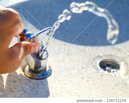 Hand operating chrome water fountain with flowing water stream in outdoor public park Hand operating chrome water fountain with flowing water stream in outdoor public park 126711259
