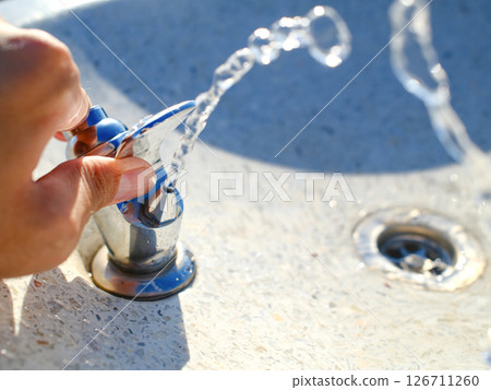 Hand operating chrome water fountain with flowing water stream in outdoor public park 126711260