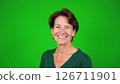 Cheerful middle-aged woman smiles warmly in studio. Portrait shot against plain green background. Attractive woman in casual green sweater. Pro, friendly expression. Mid-life beauty, casual style 126711901