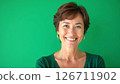 Cheerful middle-aged woman smiles warmly in studio. Portrait shot against plain green background. Attractive woman in casual green sweater. Pro, friendly expression. Mid-life beauty, casual style 126711902