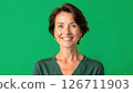 Cheerful middle-aged woman smiles warmly in studio. Portrait shot against plain green background. Attractive woman in casual green sweater. Pro, friendly expression. Mid-life beauty, casual style 126711903