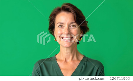 Cheerful middle-aged woman smiles warmly in studio. Portrait shot against plain green background. Attractive woman in casual green sweater. Pro, friendly expression. Mid-life beauty, casual style 126711903