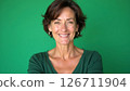 Cheerful middle-aged woman smiles warmly in studio. Portrait shot against plain green background. Attractive woman in casual green sweater. Pro, friendly expression. Mid-life beauty, casual style 126711904