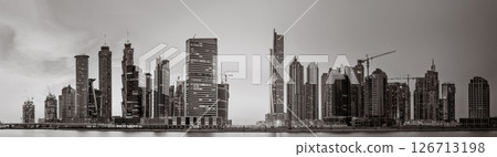Monochrome city skyline with tall skyscrapers reflecting on calm water under a cloudy sky, Dubai Marina bay UAE 126713198