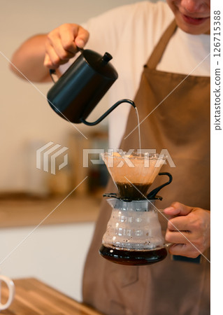 Young man in apron brewing coffee using the pour-over method in warm kitchen 126715388