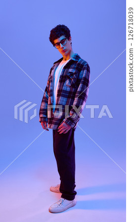 Full-body photo of teen boy in glasses standing with relaxed posture and tilted head, curious gaze creating thoughtful mood in neon against gradient background 126718039