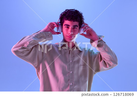 Teen boy, dressed casualwear, puts on headphones with slight smile, enjoying sound of musical immersion in neon light against gradient background Teen boy, dressed casualwear, puts on headphones with slight smile, enjoying sound of musical immersion in neon light against gradient background 126718069