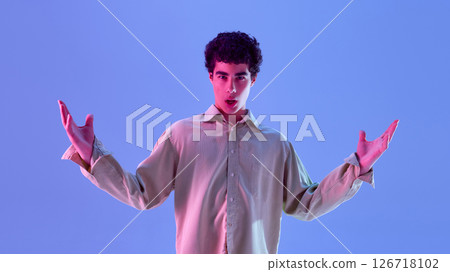 Teenage boy raising both hands with surprised expression, posing with open mouth and spreads hands wide in neon light against gradient background Teenage boy raising both hands with surprised expression, posing with open mouth and spreads hands wide in neon light against gradient background 126718102