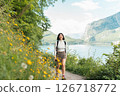 Glowing with joy, a woman strolls a nature trail wrapped in greenery, grinning as mountains and a sparkling lake unfold her perfect summer reset 126718772