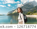 Wandering off the grid, a smiling woman walks a winding path through lush greenery, with mountain peaks and a calm lake setting the tone for mindful summer travel 126718774