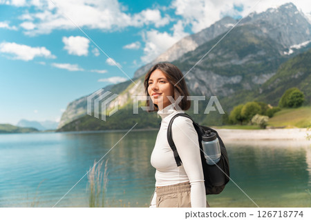 Wandering off the grid, a smiling woman walks a winding path through lush greenery, with mountain peaks and a calm lake setting the tone for mindful summer travel Wandering off the grid, a smiling woman walks a winding path through lush greenery, with mountain peaks and a calm lake setting the tone for mindful summer travel 126718774