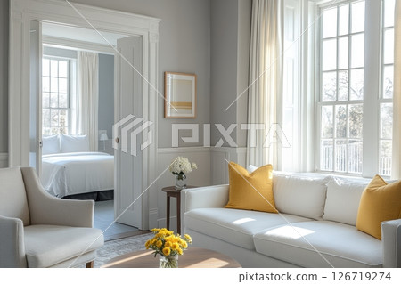 Bright modern living room interior with elegant white furniture, yellow accent pillows, large windows and classic French paneling. Natural light and fresh flowers. Sophisticated and cozy touch. 126719274