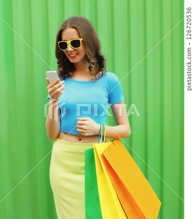 Beautiful happy young woman looking at phone with colorful shopping bags on green background 126720536
