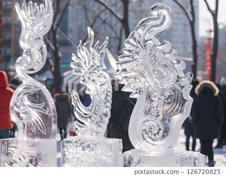 Ice sculptures in a winter festival. Intricate designs of dragons and mythical creatures carved from clear ice. Snowy ground and blurred figures in the background. 126720825