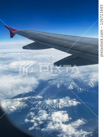 View from airplane window showing wing , blue sky and snow-covered mountain range partially covered by clouds, transportation and travel concept View from airplane window showing wing , blue sky and snow-covered mountain range partially covered by clouds, transportation and travel concept 126721685