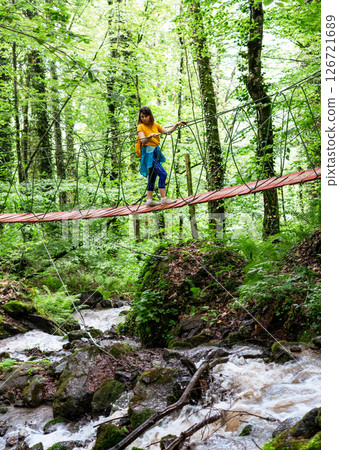 Woman hiker with yellow backpack crossing a suspension bridge in green forest over mountain stream, outdoor adventure in nature 126721689