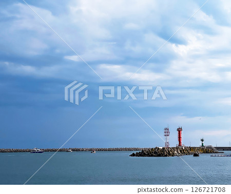 Coastal landscape showcases a lighthouse and breakwater, with boats gently floating in the water, under a sky filled with dynamic clouds, evoking tranquility and beauty 126721708