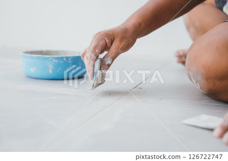 Hand gripping a rubber float tool while pressing grout into the spaces between ceramic floor tiles. Close-up showing seamless tile work. 126722747