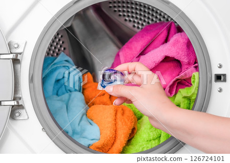 female hand holding washing capsule against background of washing machine with colored laundry female hand holding washing capsule against background of washing machine with colored laundry 126724101
