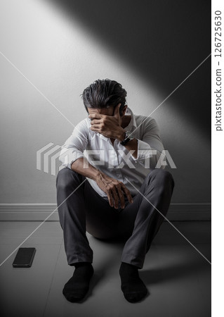 Depressed man sitting alone on the floor in dramatic lighting, covering face with hand, feeling hopeless and emotionally overwhelmed 126725630