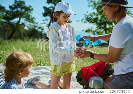A mother pours a drink for her children during a sunny picnic in the mountains, emphasizing family bonding, outdoor leisure, and the beauty of nature. 126725963