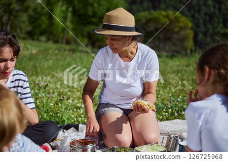 A family gathers for a joyful summer picnic in a scenic mountain meadow. The mother delights in a taco while surrounded by children, embracing the beauty of nature and family bonds. A family gathers for a joyful summer picnic in a scenic mountain meadow. The mother delights in a taco while surrounded by children, embracing the beauty of nature and family bonds. 126725968