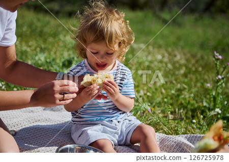 A child enjoys eating corn while spending quality time with their mother outdoors during a sunny picnic in a serene natural environment. 126725978