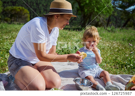 A woman and her young child share a joyful picnic scene outdoors. The warm and sunny day enhances the serene mountain backdrop, emphasizing family bonding and love for nature. A woman and her young child share a joyful picnic scene outdoors. The warm and sunny day enhances the serene mountain backdrop, emphasizing family bonding and love for nature. 126725987