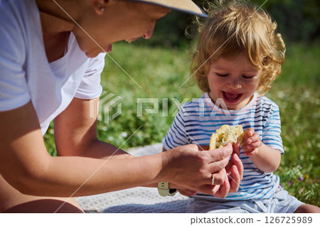 A mother and her young child sharing a joyful outdoor picnic experience on a sunny summer day, surrounded by nature, bonding and enjoying a tasty meal. 126725989