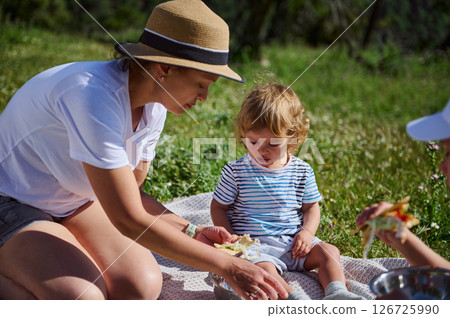 A charming family moment as a mother spends joyful time with her child during a picnic in the scenic mountains, surrounded by natural beauty under warm summer sunshine. 126725990