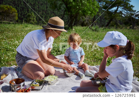 A mother and her children share a delightful outdoor picnic on a sunny day in a scenic mountain landscape. 126725991