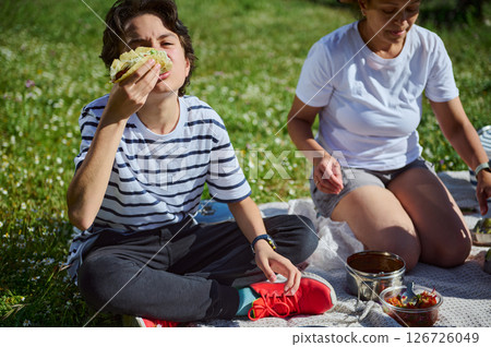 A family spends a beautiful sunny day enjoying a picnic in a lush green outdoor setting. A child eats a taco with a joyful expression while the mother assists with picnic arrangements. 126726049