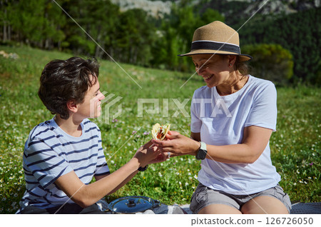 Mother and son spending quality time on a mountainside, sharing food, sunshine, and laughter during a joyful summer picnic amid scenic nature. 126726050