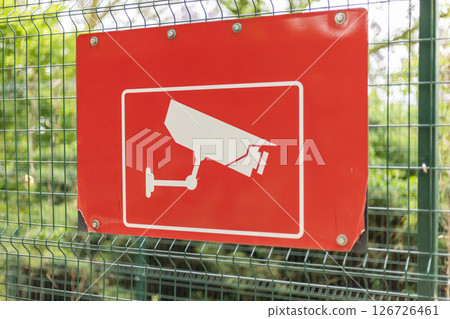A bright red sign featuring a white CCTV camera graphic is mounted on a fence amidst lush greenery. The sign serves to inform about surveillance measures in the area 126726461