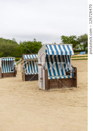 Colorful beach chairs with blue and white stripes are set up on a sandy beach. The overcast sky creates a calm atmosphere, with greenery in the distance Colorful beach chairs with blue and white stripes are set up on a sandy beach. The overcast sky creates a calm atmosphere, with greenery in the distance 126726470