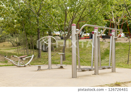 A well-maintained outdoor fitness area features multiple exercise stations designed for public use. The greenery surrounds the equipment, inviting community fitness activities A well-maintained outdoor fitness area features multiple exercise stations designed for public use. The greenery surrounds the equipment, inviting community fitness activities 126726481