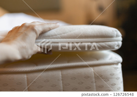 A hand gently lifts a comfort layer from a mattress in a softly lit bedroom. The bedding appears fresh and inviting, ready for relaxation and restful sleep A hand gently lifts a comfort layer from a mattress in a softly lit bedroom. The bedding appears fresh and inviting, ready for relaxation and restful sleep 126726548