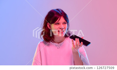 Girl holding phone and talking with funny expression under soft neon background showing casual interaction Girl holding phone and talking with funny expression under soft neon background showing casual interaction 126727509