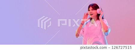 Teen girl with headphones joyfully talking waving hands showing excitement under vibrant light against gradient background with negative space. Banner 126727515