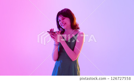 Laughing young girl, teenager in green dress holding smartphone and enjoying moment under pink violet neon lighting against gradient background. 126727558