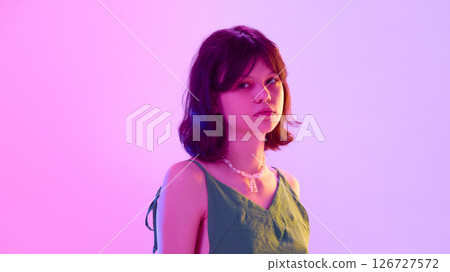 Stylish teen girl with piercings wearing green dress and pearl necklace looking calm under purple pink lighting against gradient background Stylish teen girl with piercings wearing green dress and pearl necklace looking calm under purple pink lighting against gradient background 126727572