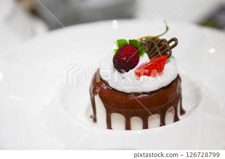On a white plate, a small cake doused with chocolate glaze and decorated with berries. On a white plate, a small cake doused with chocolate glaze and decorated with berries. 126728799