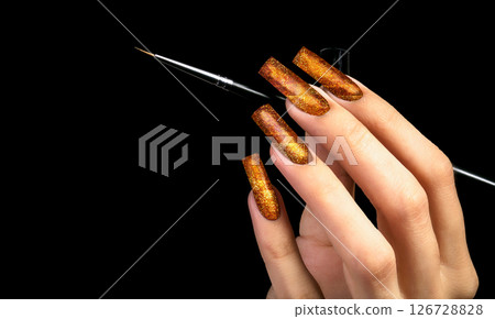 Hand with a beautiful gold manicure and a brush on a black background. Nail design. Extended nails. Hand with a beautiful gold manicure and a brush on a black background. Nail design. Extended nails. 126728828