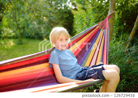 A cute blonde white preteen boy is having fun in the summer in a colorful hammock in the backyard or on the outdoor playground. A child is resting and swinging in a hammock. 126729438