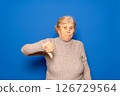 Caucasian woman in her 70s wearing a dark beige sweater with her thumb down in a sign of disgust and disapproval, isolated on blue studio background. 126729564